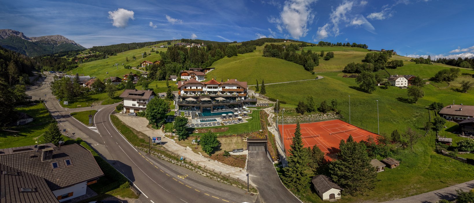 Hotel + Plan de Corones + Olang = mountain biking, climbing, tennis View of a hotel with pool and tennis court in green mountain landscape