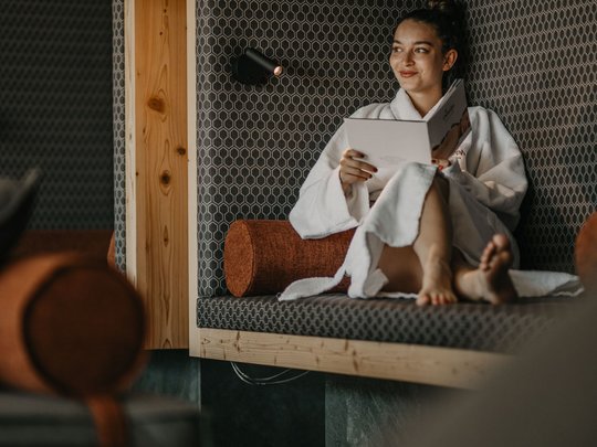 Hotel al Plan de Corones con spa: vacanza à la Christoph Donna in accappatoio si rilassa su divano leggendo un opuscolo in ambiente moderno