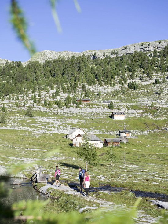 Der Kronplatz aus nächster Nähe: Ihr Aktivhotel in Südtirol Wanderer gehen auf einem Pfad in einer bergigen Landschaft mit Häusern und Wald