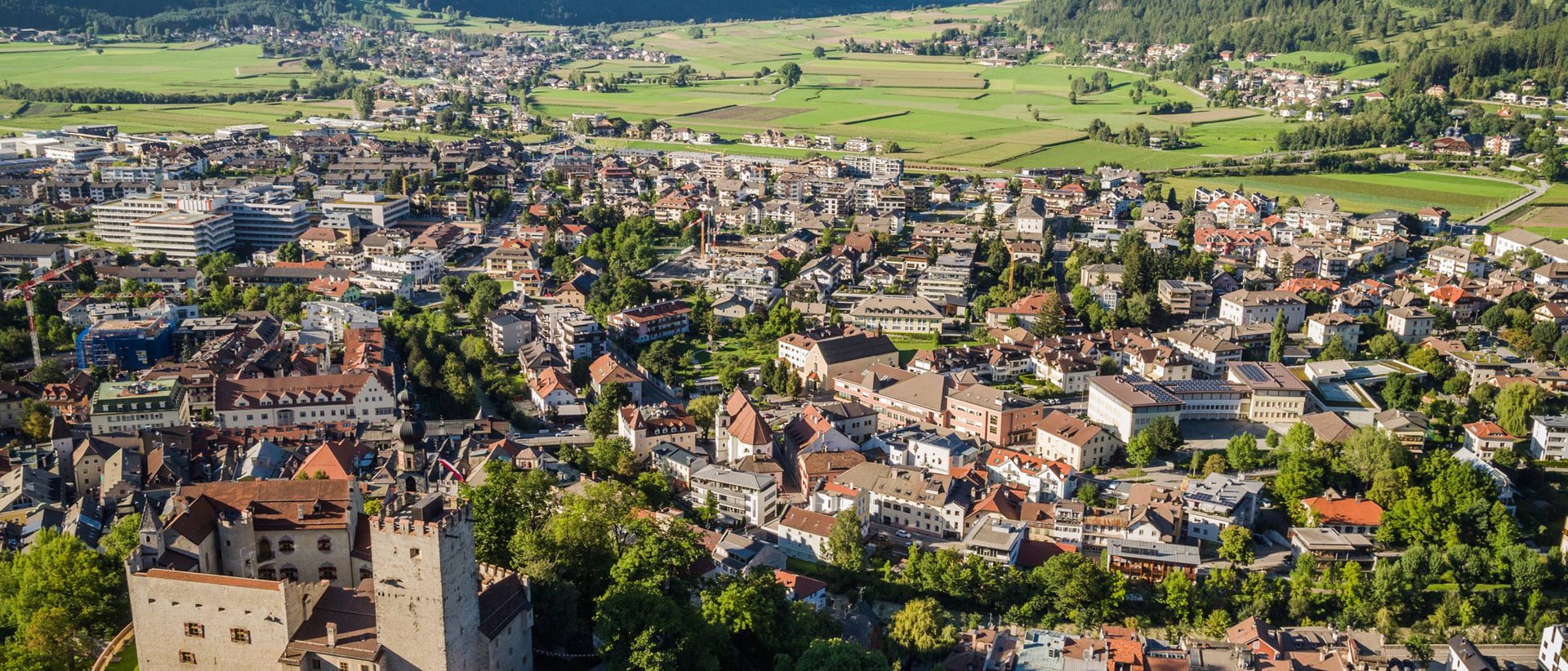 Gemütlicher Stadtbummel durch Bruneck Luftaufnahme einer Stadt in einem Tal mit einer Burg und Bergen im Hintergrund