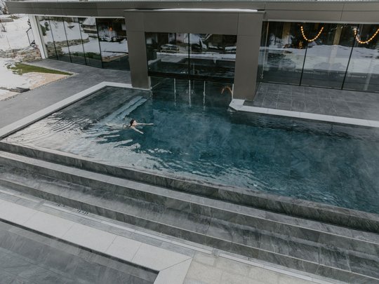 Hotel in Val Pusteria con piscina Persona che nuota in piscina esterna moderna accanto a edificio con neve invernale.