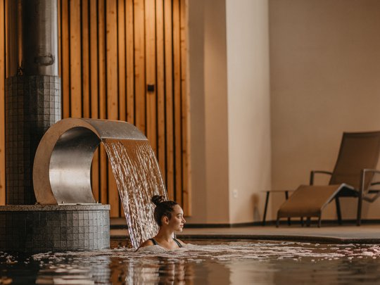 Le novità del nostro hotel a 4 stelle tra le Dolomiti Donna rilassata in piscina interna sotto cascata con parete in legno e sdraio