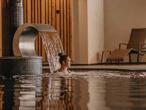 Aufregende Neuigkeiten aus unserem Hotel in den Dolomiten Frau entspannt im Innenpool unter Wasserfall mit Holzwand und Liegestuhl