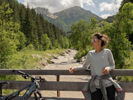 Hotel + Plan de Corones + Olang = mountain biking, climbing, tennis Woman leaning on bridge railing with bike in front of mountain landscape and river