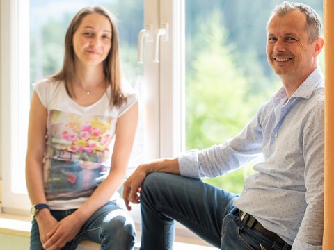 Regionale, alpine Küche im Hotel am Kronplatz in Südtirol Zwei lächelnde Personen sitzen am Fenster mit Vorhang im Hintergrund