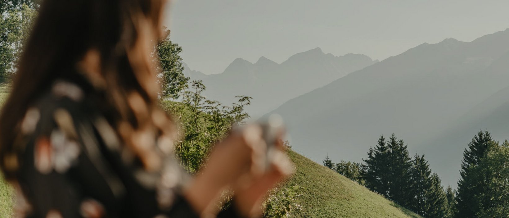 4-Sterne-Hotel am Kronplatz: Ihr perfektes Urlaubszuhause Frau mit Tasse auf Balkon blickt auf grüne Hügel und Berge bei klarem Himmel