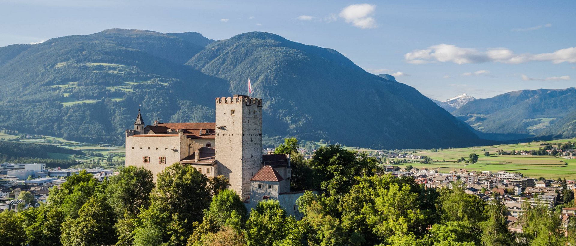 Zu Besuch in Bruneck Burg auf bewaldetem Hügel mit Bergen und Stadt im Hintergrund