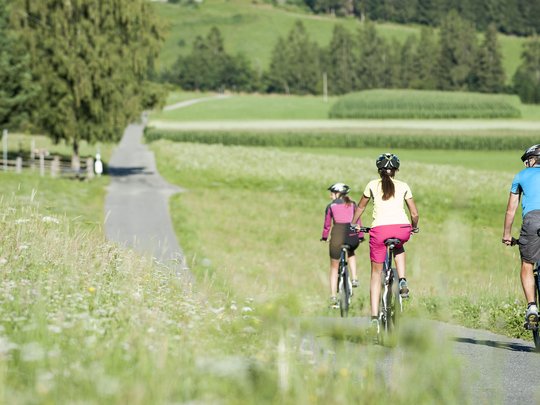Neuigkeiten unseres Hotels nahe dem Pragser Wildsee Drei Personen fahren Fahrrad auf einem Landweg durch grüne Wiesen