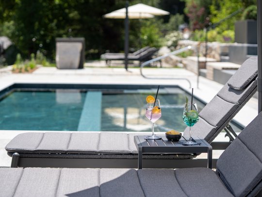Le novità del nostro hotel a 4 stelle tra le Dolomiti Due lettini grigi con bevande accanto a una piscina in giardino