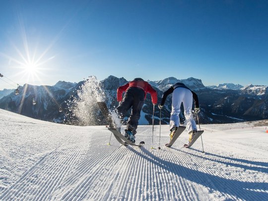 Unforgettable skiing holiday on Plan de Corones/Kronplatz Two skiers starting on a sunny groomed ski slope with mountains in the background