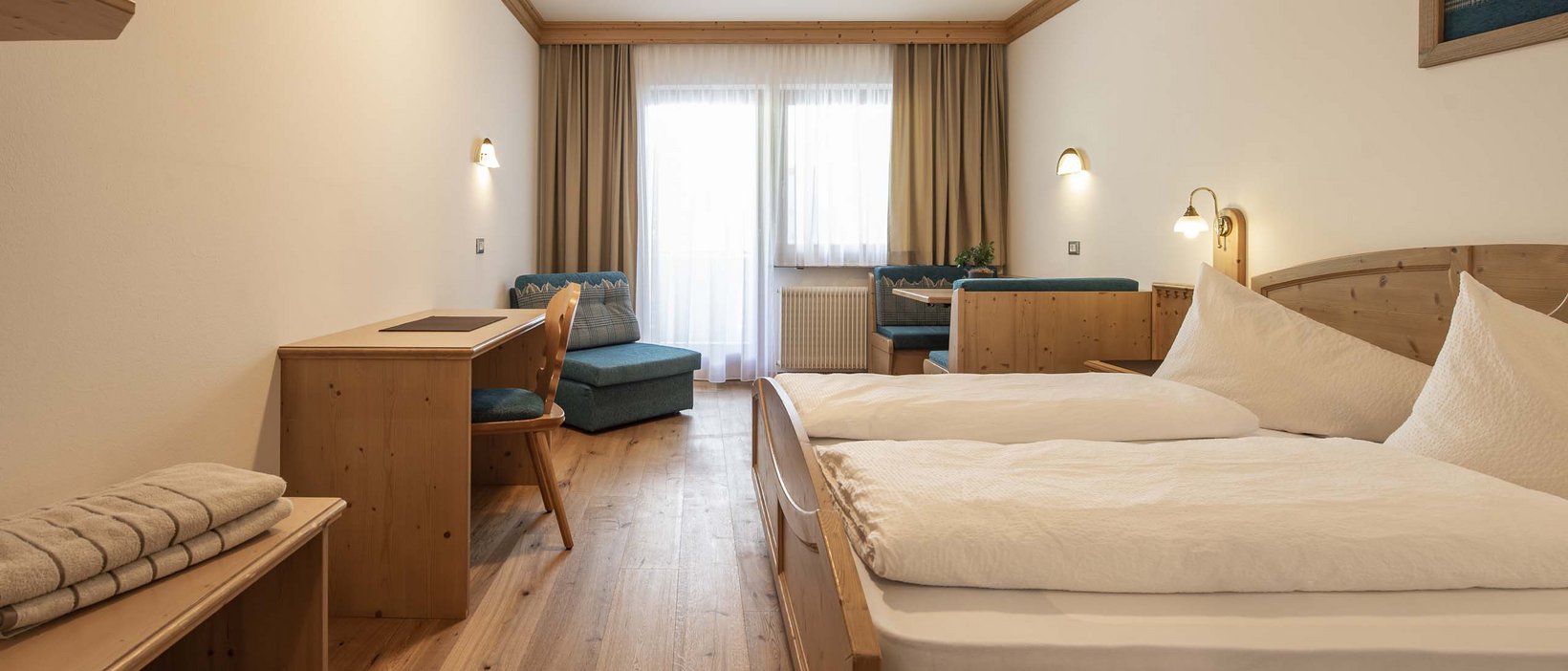 A warm welcome to our hotel in Olang, at Plan de Corones/Kronplatz Bright bedroom with wooden furniture, double bed, and seating area