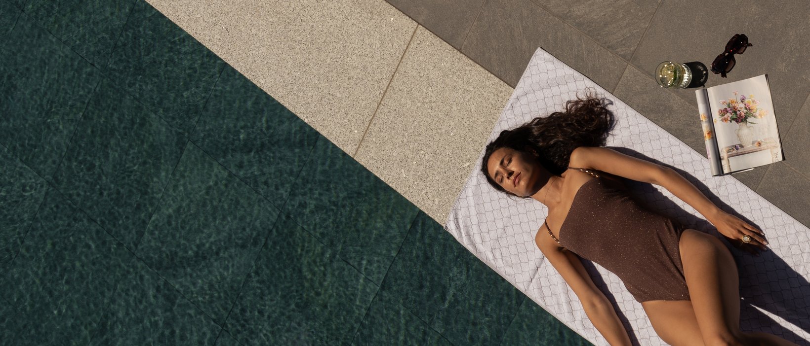 Una giornata di relax Donna sdraiata su asciugamano accanto alla piscina con libro, bevanda e occhiali da sole