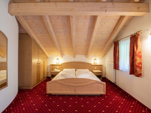 Info e prezzi del nostro hotel benessere nelle Dolomiti Camera da letto accogliente con soffitto in legno, letto matrimoniale e moquette rossa
