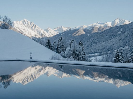 4-Sterne-Hotel am Kronplatz: Ihr perfektes Urlaubszuhause Schneebedeckte Berge spiegeln sich in einem ruhigen See unter blauem Himmel