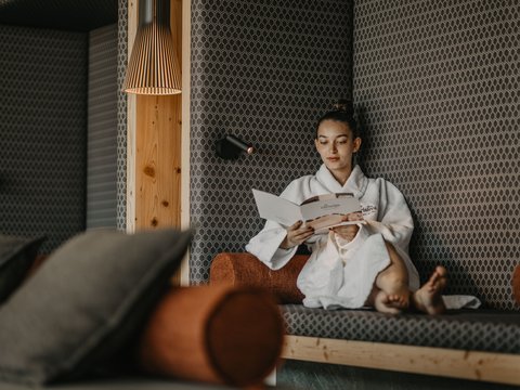 Baden im Pragser See? Hotel mit Pool? Brrr, ab in den Pool! Frau liest entspannt in einem Bademantel auf einer Couch in modernem Raum