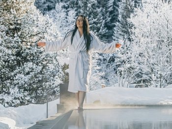 Aufregende Neuigkeiten aus unserem Hotel in den Dolomiten Frau im Bademantel am Rand eines dampfenden Pools mit schneebedecktem Wald