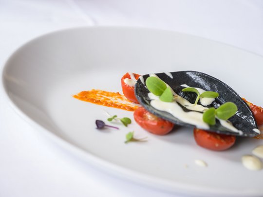 Regionale, alpine Küche im Hotel am Kronplatz in Südtirol Schwarze Ravioli mit Tomaten, grünen Blättern und weißer Soße auf weißem Teller